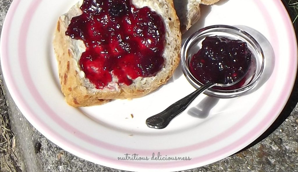 Sliced of Walnut and Gruyere loaf with butter and blackcurrant jam. Small pot of jam with spoon beside it.