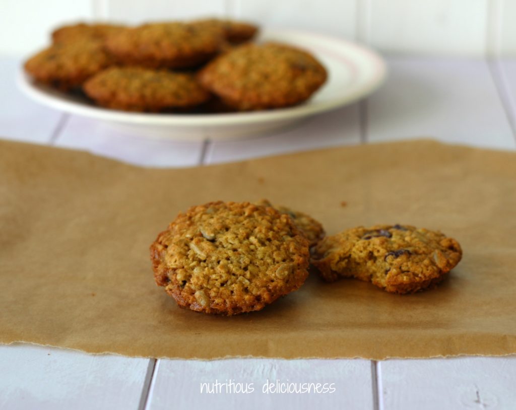 3 seedy fruity cookies on a piece of brown greaseproof paper with a white plate in the background with a pile of cookies on.