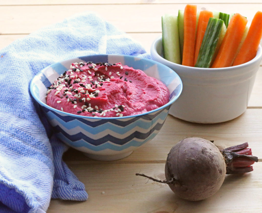 Beetroot Hummus sprinkled with black and white sesame seeds in a blue patterned bowl with carrot and courgette batons in a white pot. Two small beets on the wooden board.