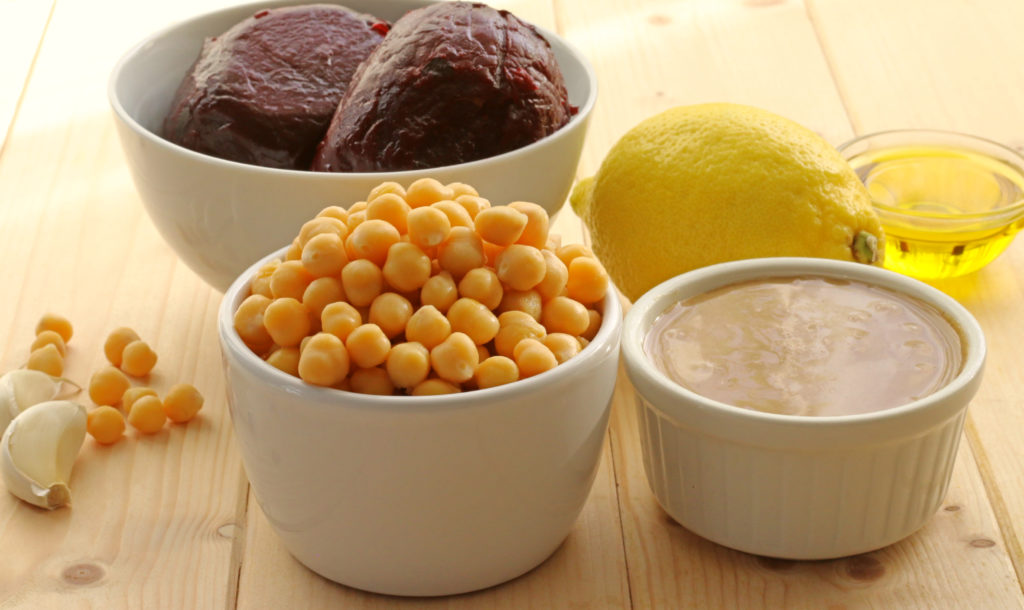 Two roasted beetroots in white bowl. In front of them, white pots filled with chickpeas (garbanzos), Tahini and olive oil.