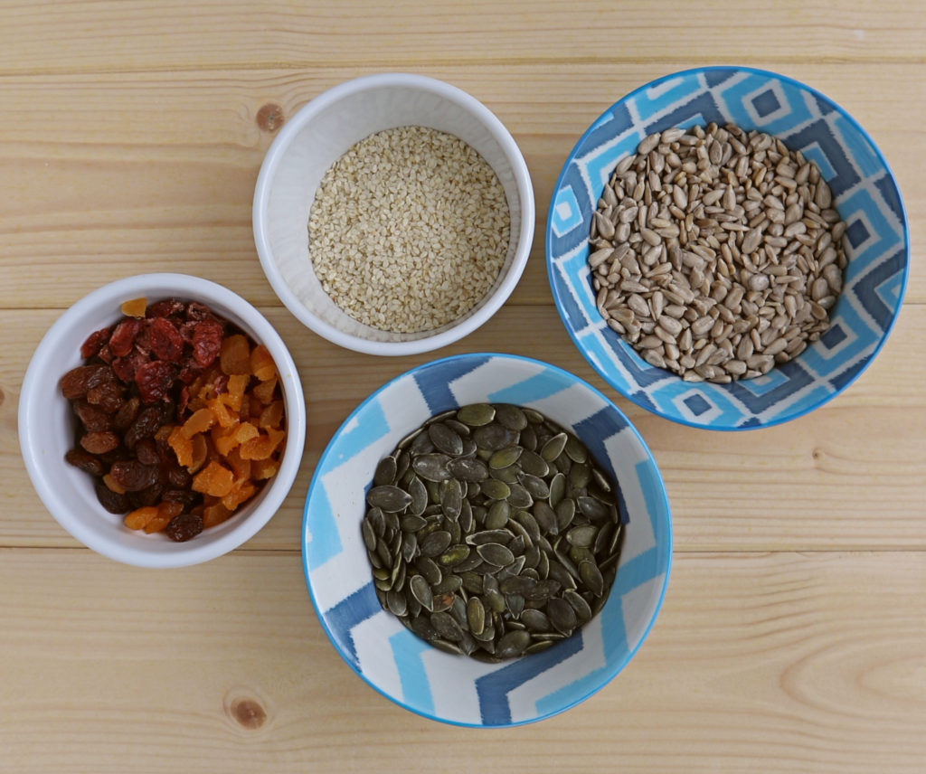granola ingredients sunflower, pumpkin and sesame seeds and dried fruit in bowls on a wooden surface.