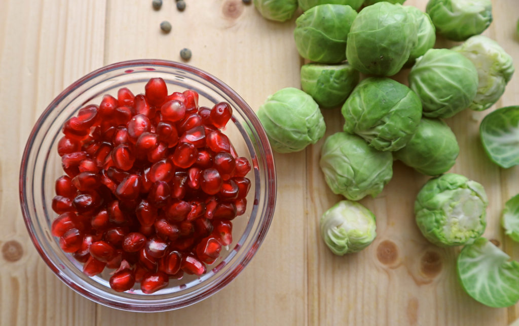 Pomegranate seeds in a glass bowl with some brussel sprouts to the side.