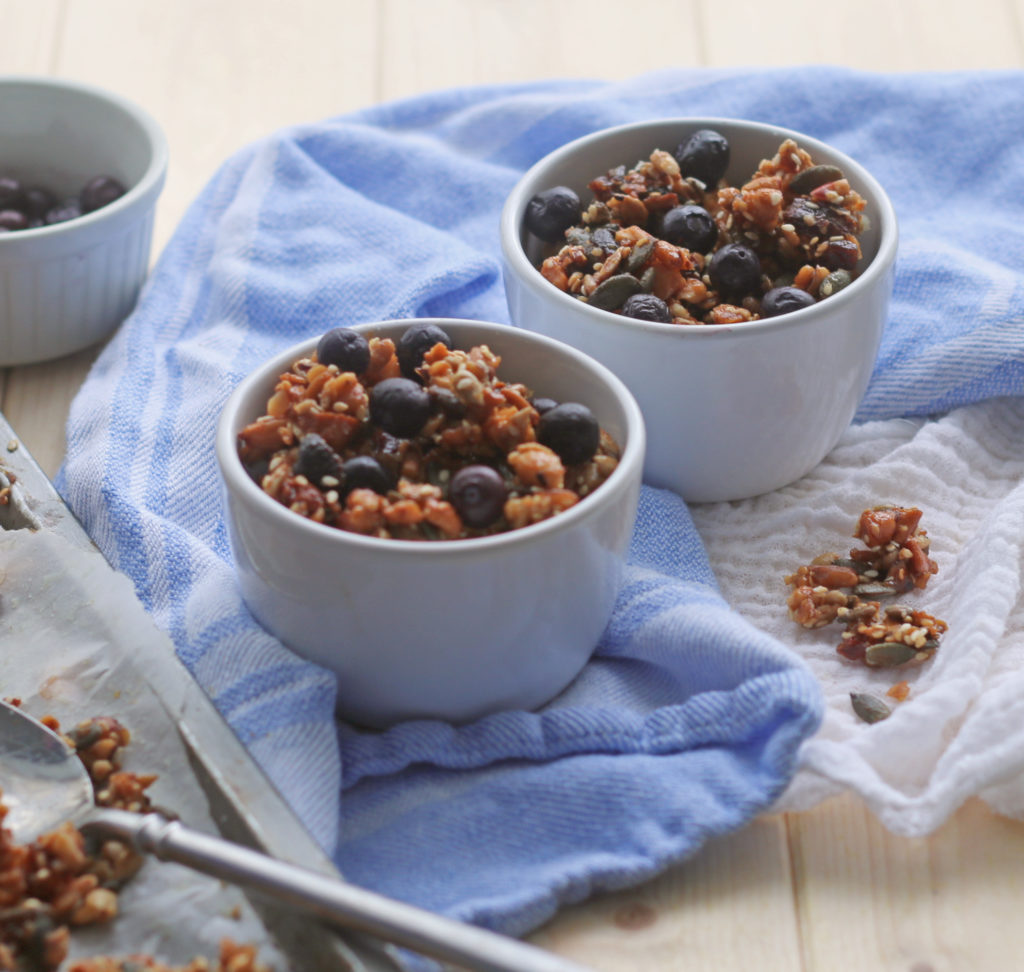 breakfast of no-grain gluten-free granola in small white bowls on a wooden surface and blue tea towel