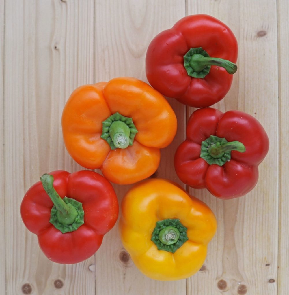 3 red peppers, 1 orange pepper and 1 yellow pepper on a wooden board.