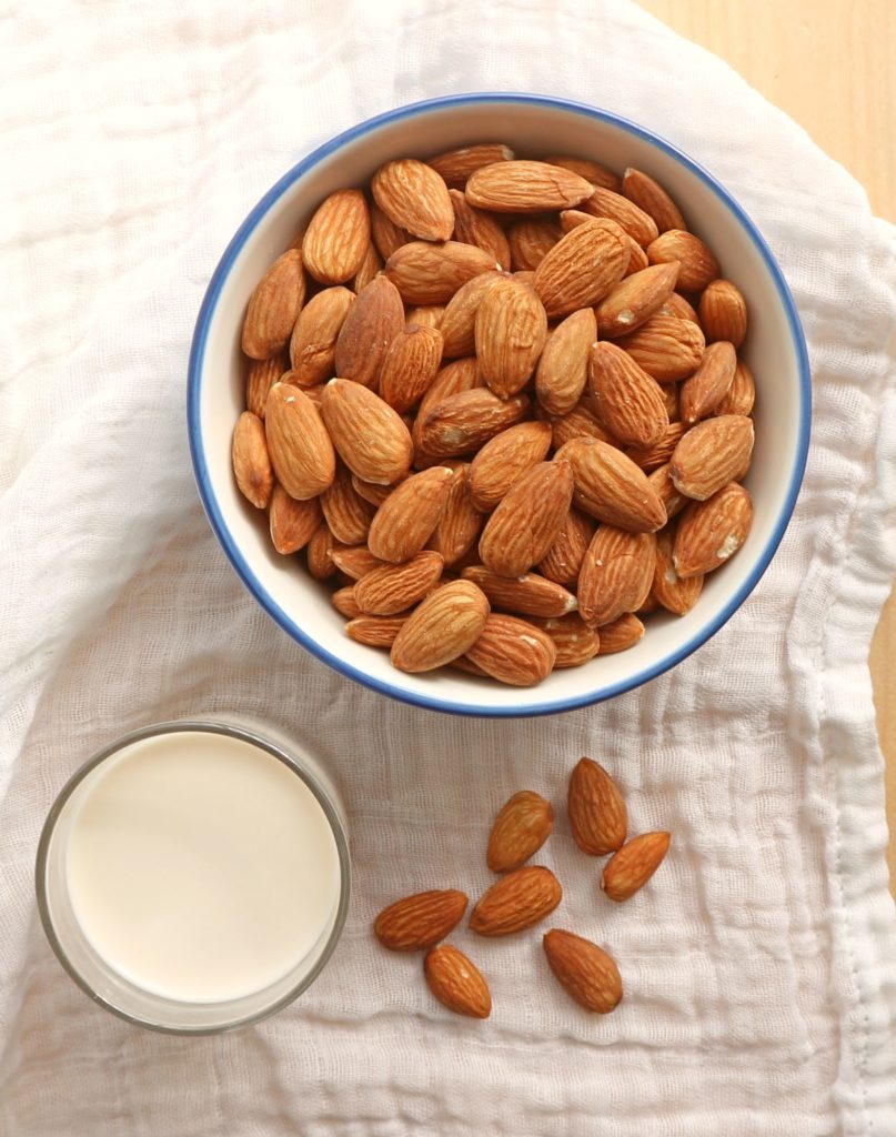 Raw almonds in a white bowl with almond milk in a glass.