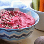 Beetroot Hummus sprinkled with black and white sesame seeds in a blue patterned bowl with carrot and courgette batons in a white pot. Two small beets on the wooden board.