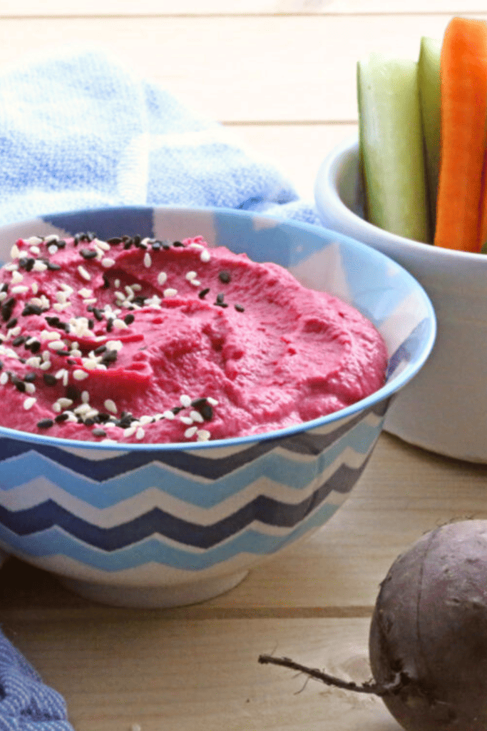 Beetroot Hummus sprinkled with black and white sesame seeds in a blue patterned bowl with carrot and courgette batons in a white pot. Two small beets on the wooden board.