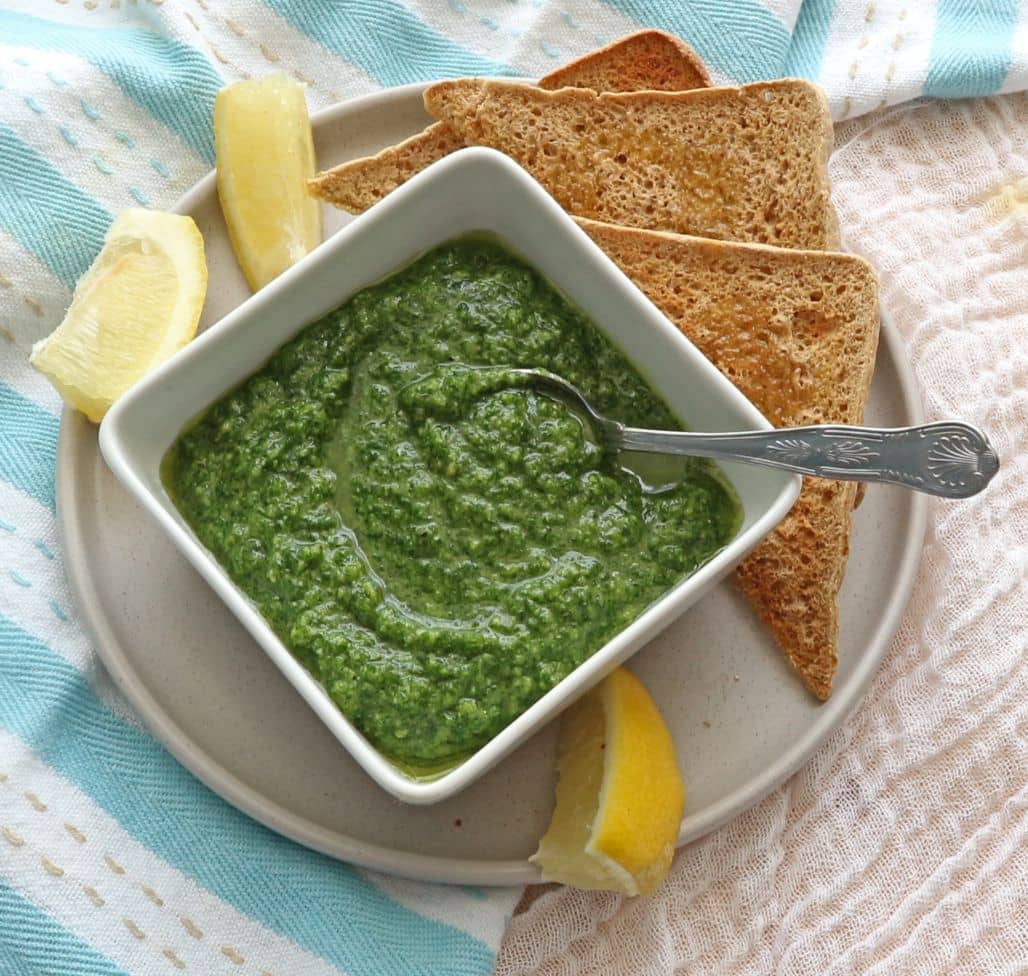 Parsley Pesto in a white bowl on a beige plate with slices of toast and lemon wedges.