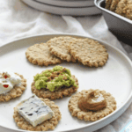 Oatcakes with various toppings on a light grey plate with oatcakes in a cookie tin.