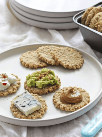 Oatcakes with various toppings on a light grey plate with oatcakes in a cookie tin.