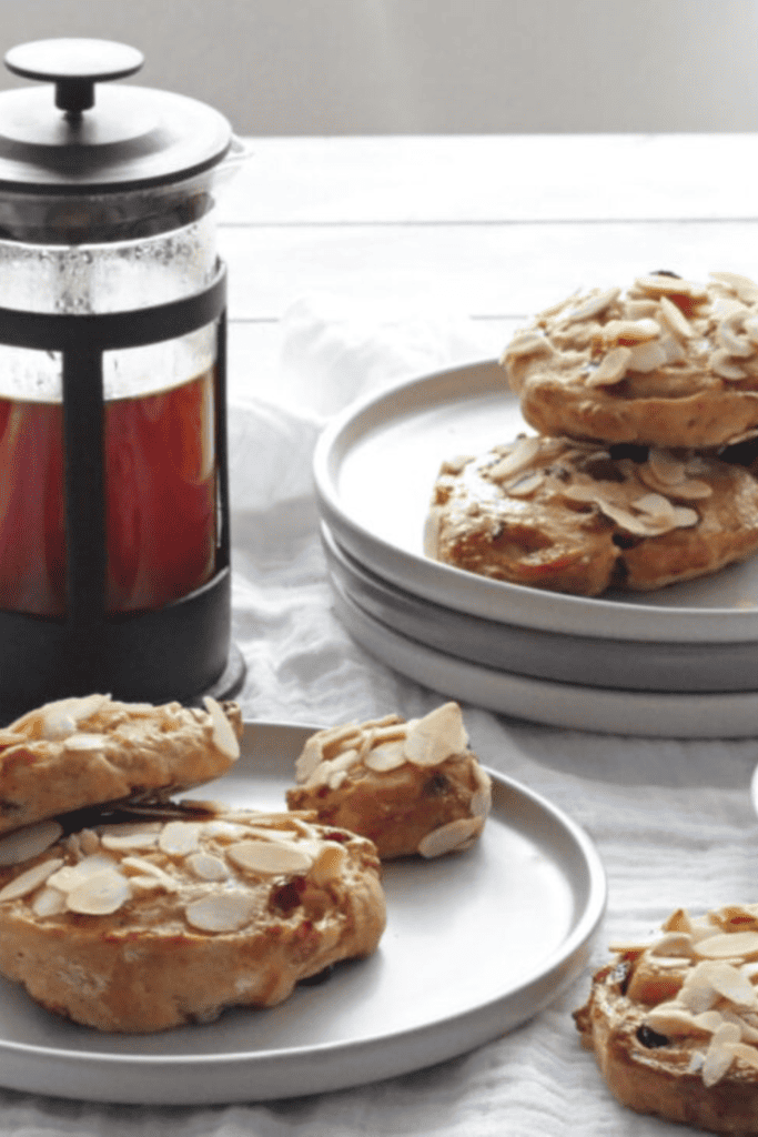 Two Stollen Pastries on a grey plate with one on the side. Plus a pile of plates with 3 Stollen Pastries on. Cafetiere full of fresh coffee and a coffee cup and saucer with black coffee.