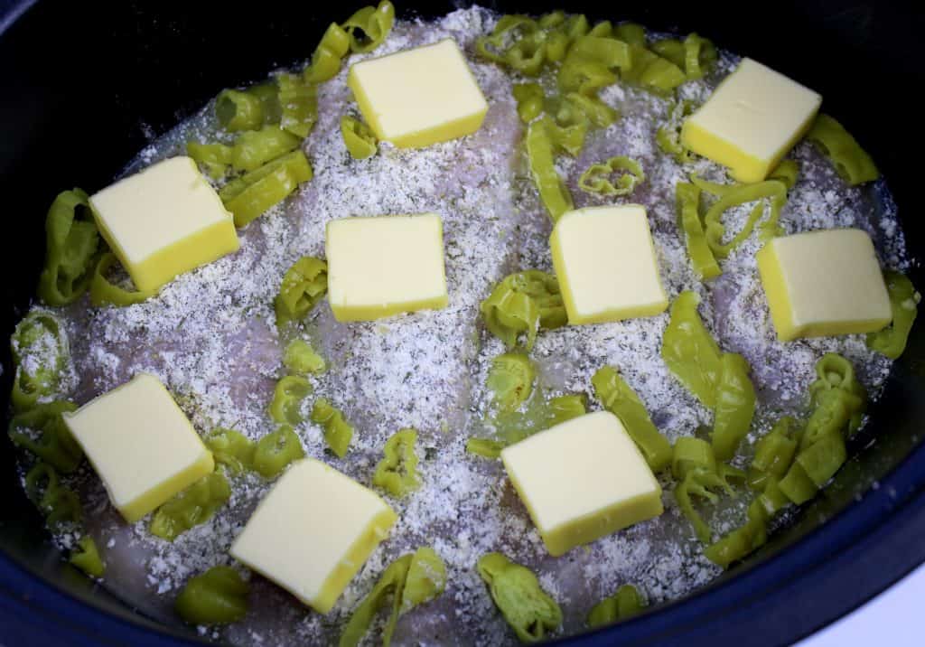 Mississippi chicken uncooked in slow cooker insert with pads of butter on top