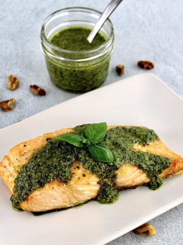 piece of salmon on beige plate with pesto sauce and basil leaf on top with basil in open jar in background