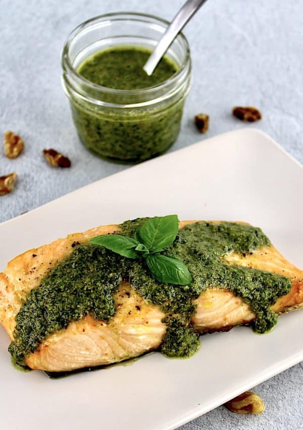 piece of salmon on beige plate with pesto sauce and basil leaf on top with basil in open jar in background
