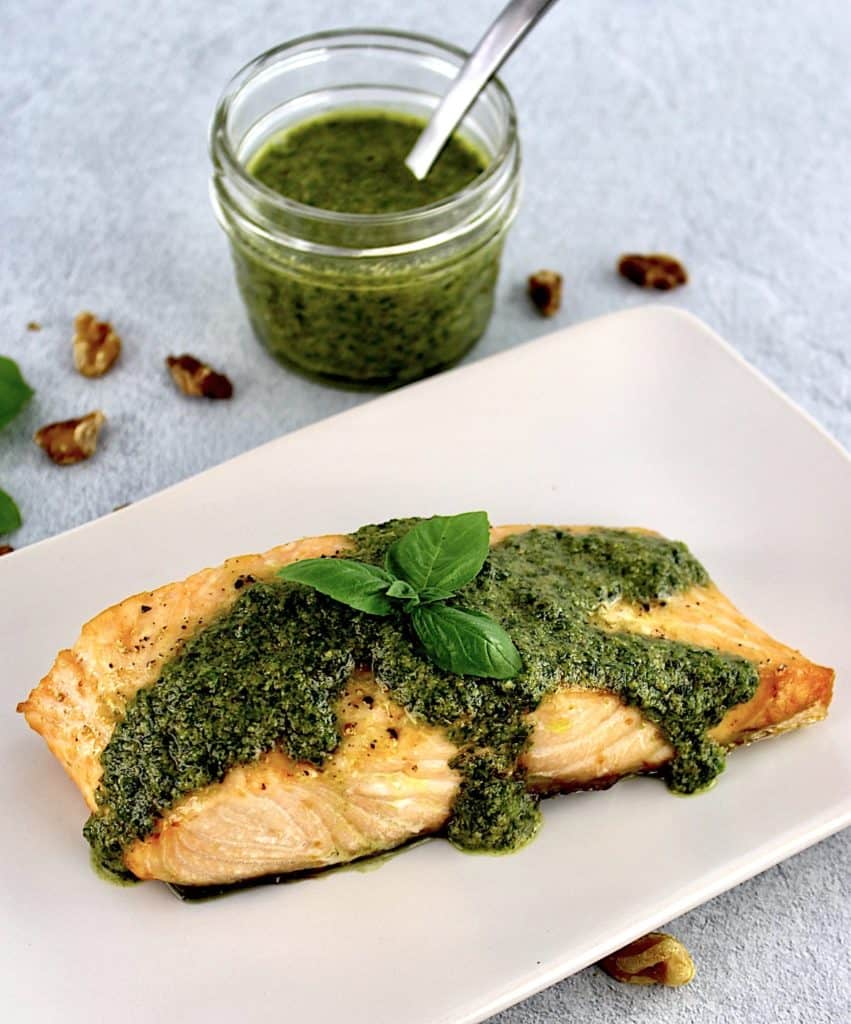 piece of salmon on beige plate with pesto sauce and basil leaf on top with basil in open jar in background