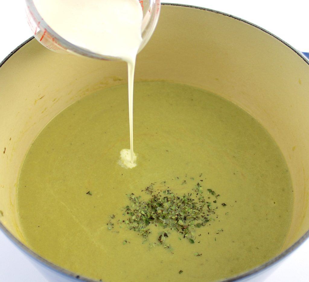 Cream of Asparagus Soup in pot with chopped thyme and cream being poured in