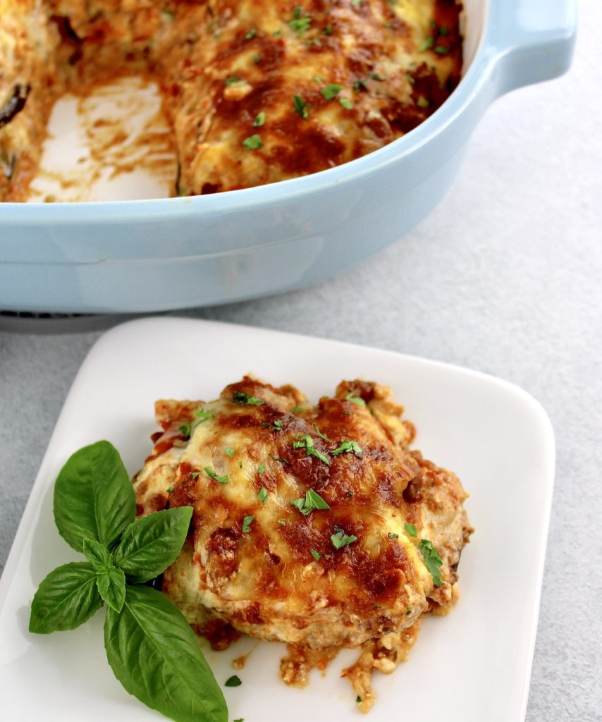 slice of Eggplant Lasagna on white dish with fresh basil on side and casserole in background