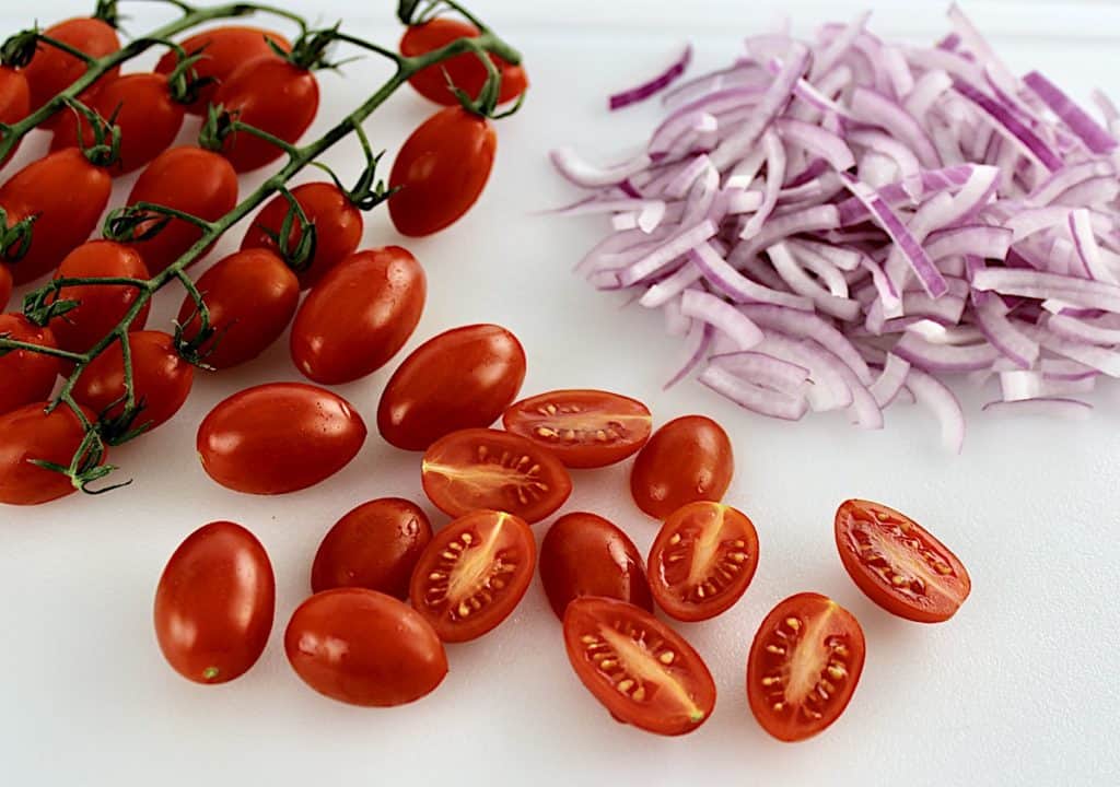 sliced tomatoes and red onions on white cutting board vine tomatoes in back