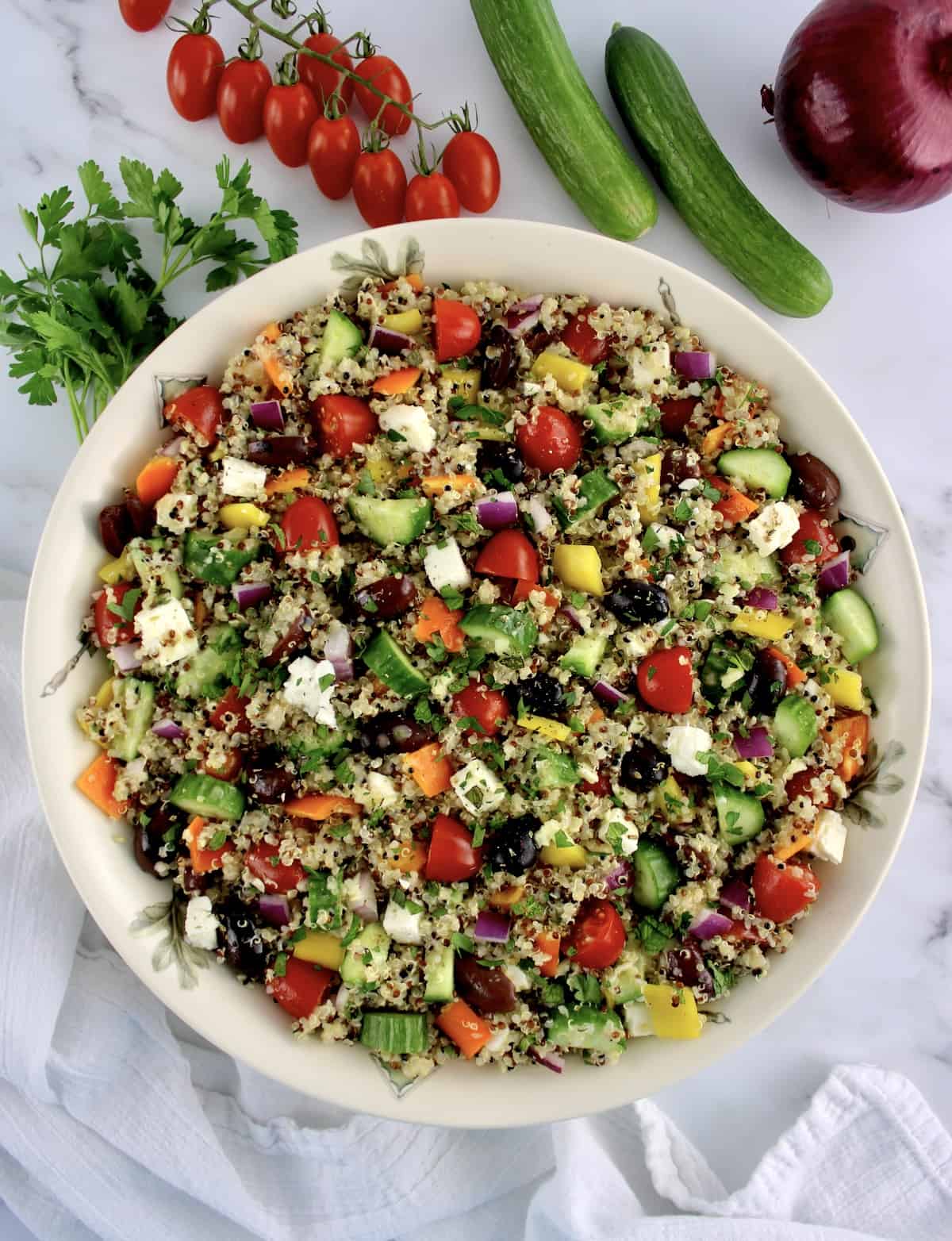 overhead view of Greek Quinoa Salad in beige bowl with parsley tomatoes and cucumbers in background