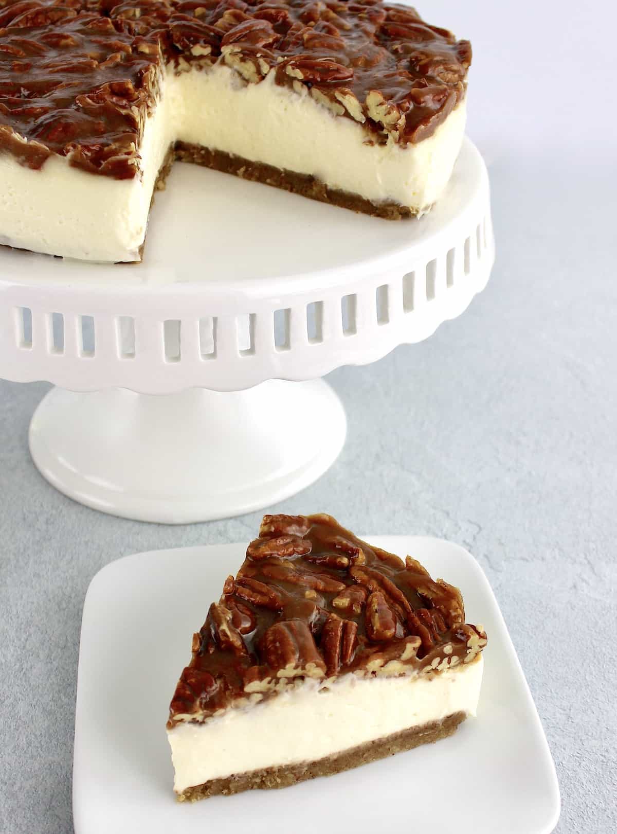 slice of pecan pie cheesecake on white plate with the rest on a white cake stand in background