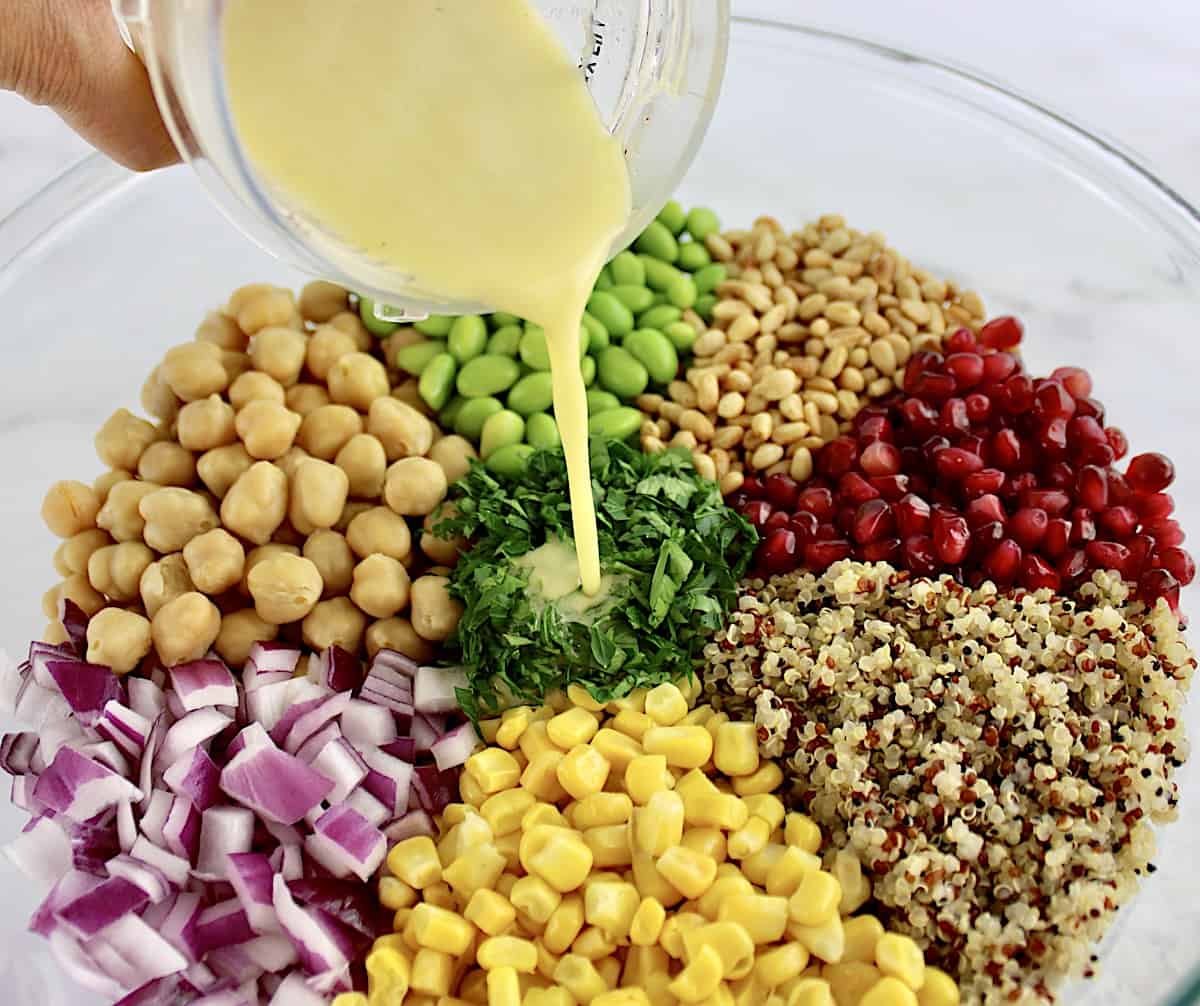 quinoa, edamame, corn, red onion, chickpeas, pomegranate seeds and parsley in glass bowl in sections with dressing being poured over top