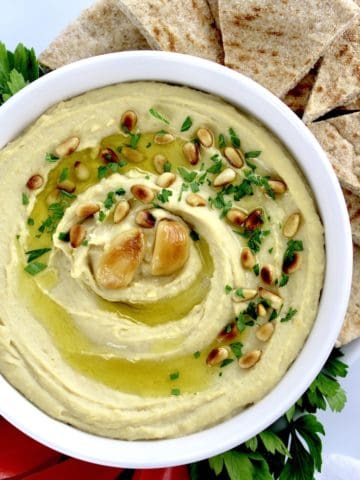 Roasted Garlic Hummus in white bowl with pine nuts, olive oil and roasted garlic on top with pita wedges on side