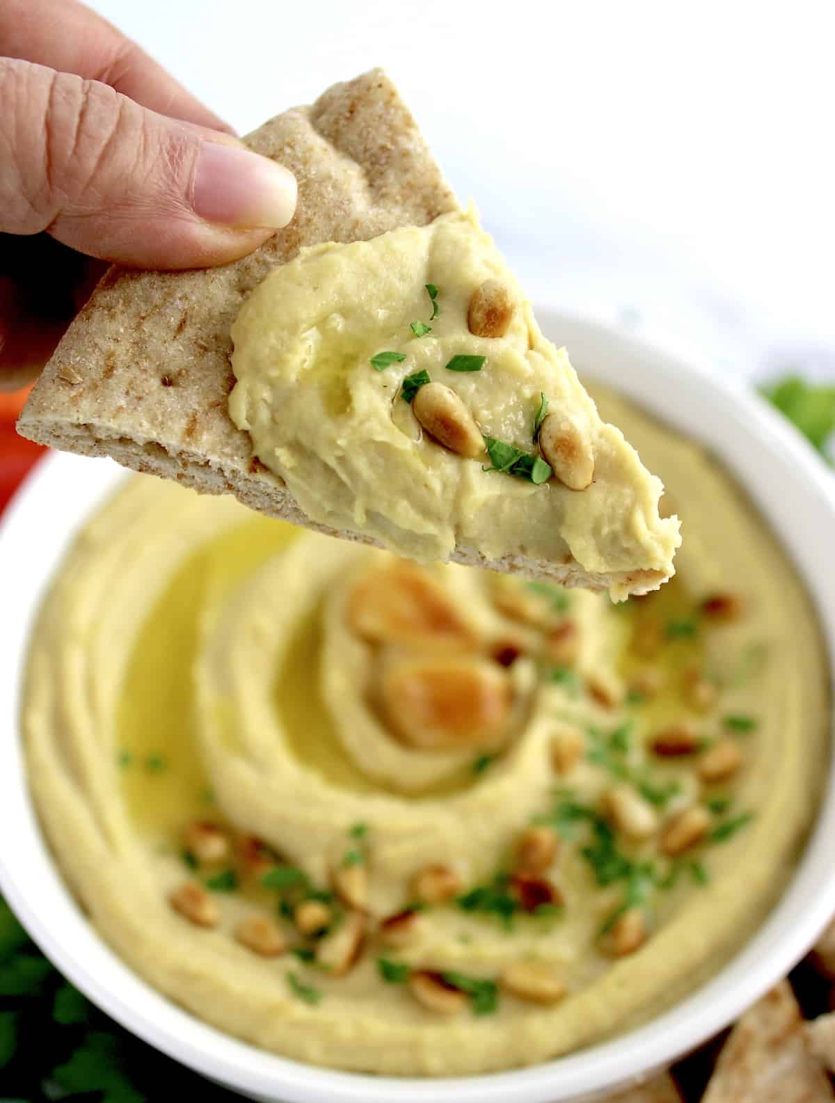 holding up pita wedge with hummus and pine nuts on top held up over bowl of hummus