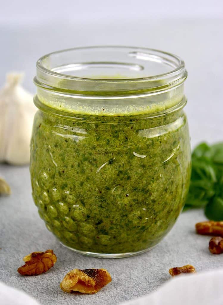 side view of Walnut Pesto in glass jar with garlic and parsley in background
