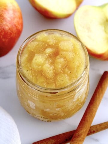 Homemade Applesauce in open glass jar with fresh apples in background with 2 cinnamon sticks