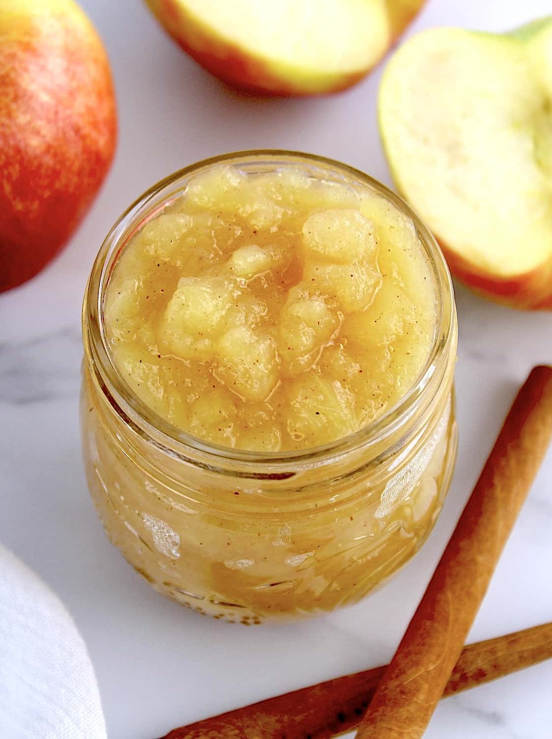 Homemade Applesauce in open glass jar with fresh apples in background with 2 cinnamon sticks