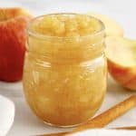 side view of Homemade Applesauce in open glass jar with cinnamon sticks and apples in background