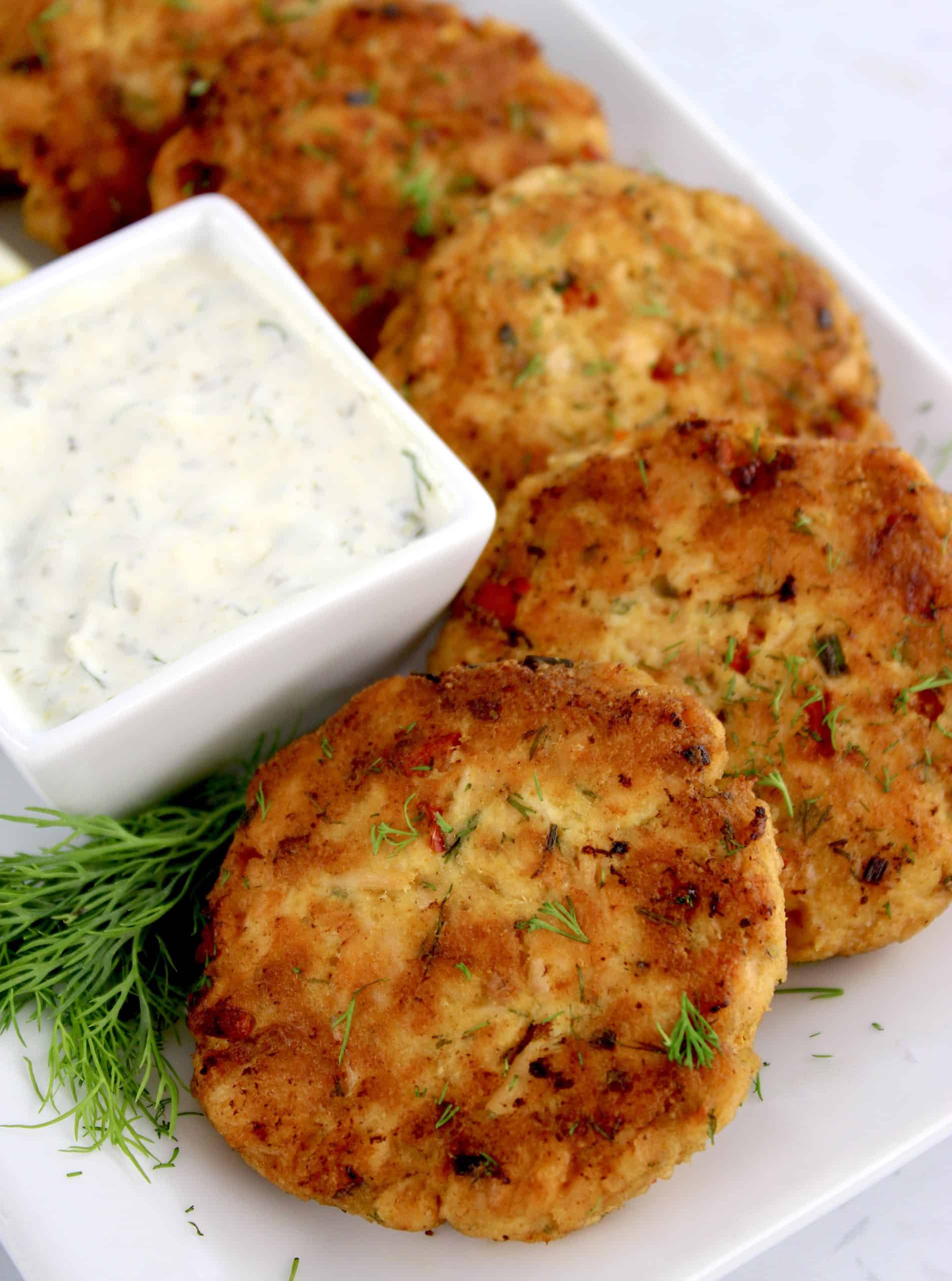 Salmon Patties lined up on white plate with fresh dill and dipping sauce on side
