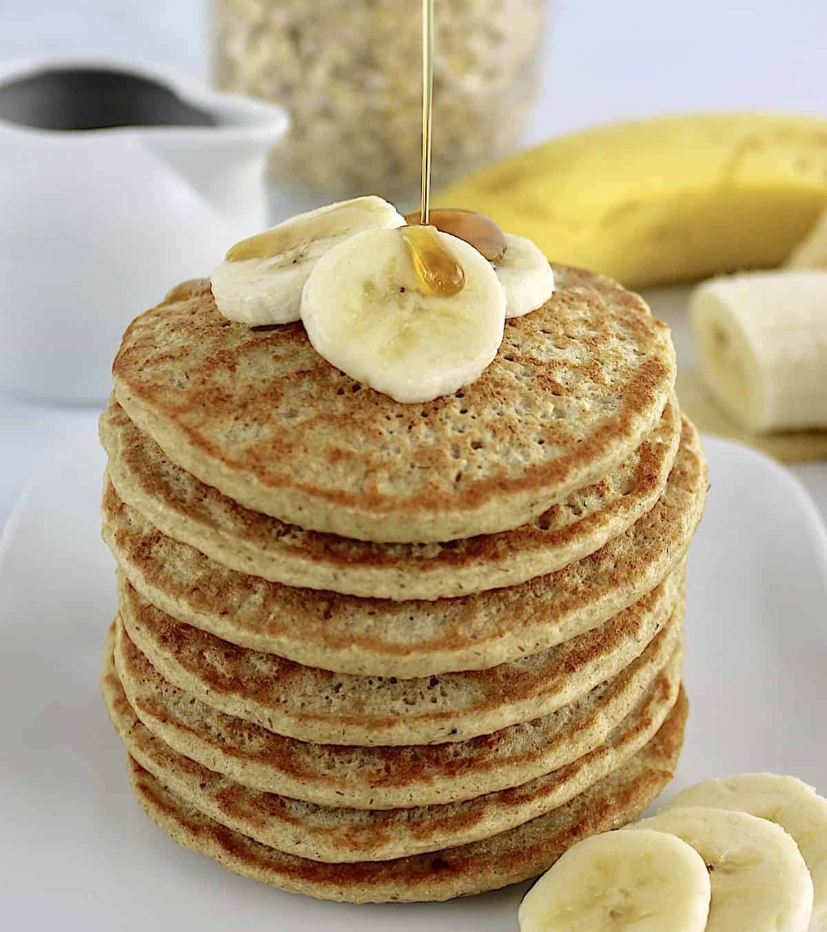 Banana Oat Flour Pancakes stack with sliced bananas on top and syrup being poured over top