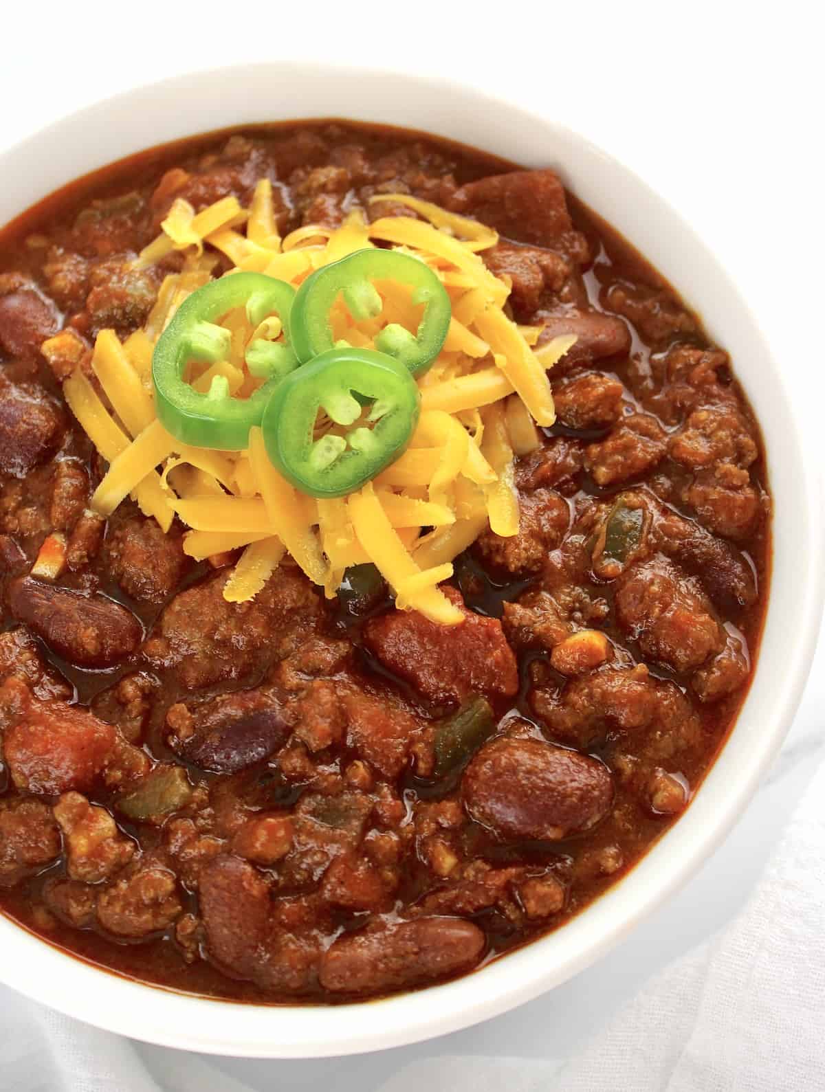 Best Homemade Chili in white bowl with shredded cheese and sliced jalapeno