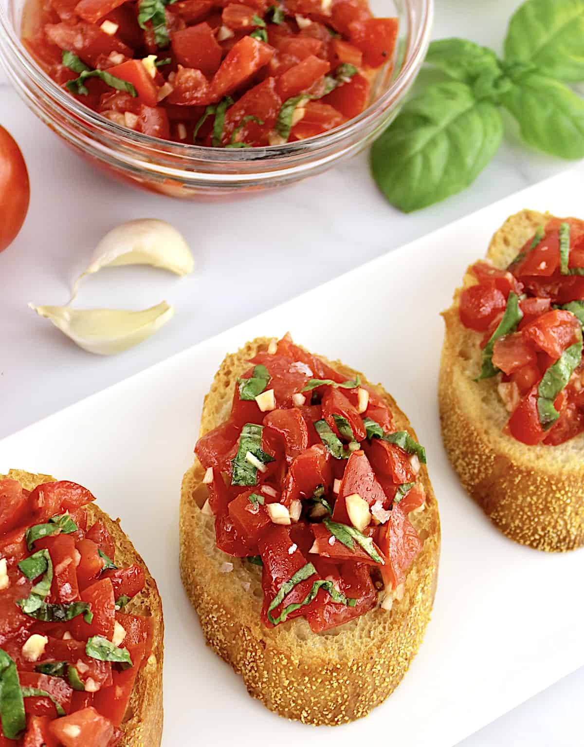 3 slices of Easy Bruschetta on white plate with tomatoes in background