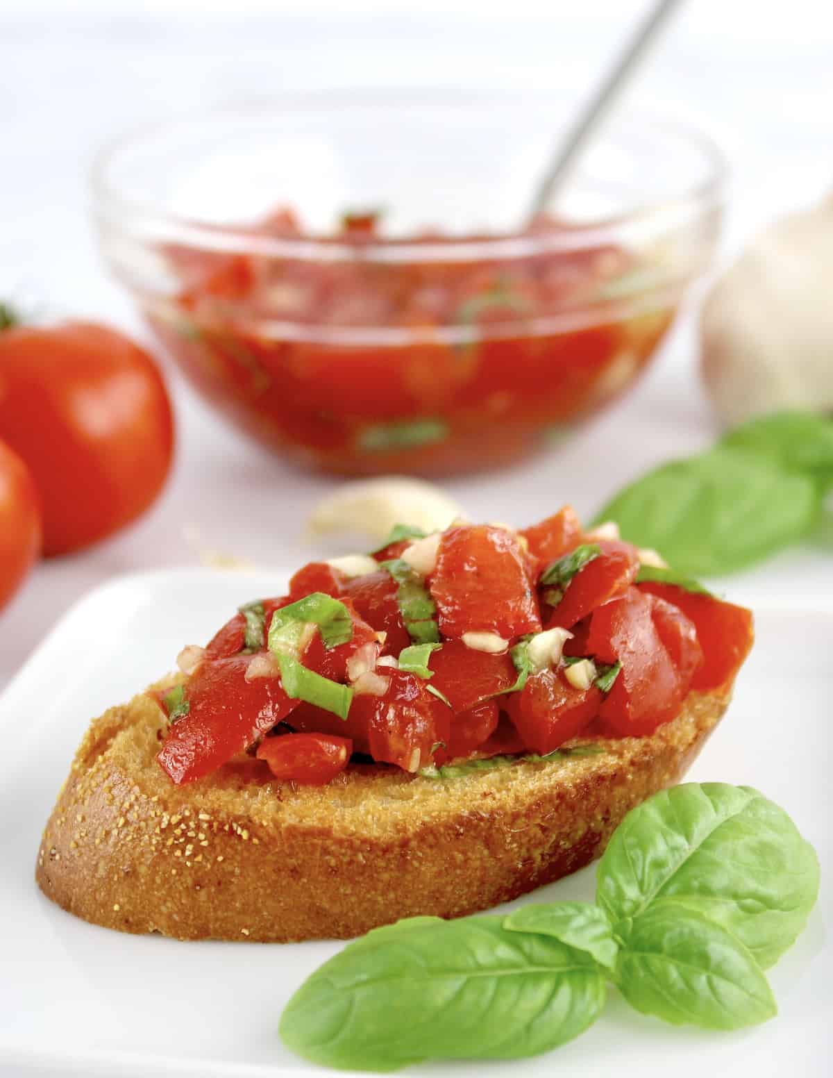 Easy Bruschetta slice on white plate with fresh basil and tomatoes in glass bowl in background