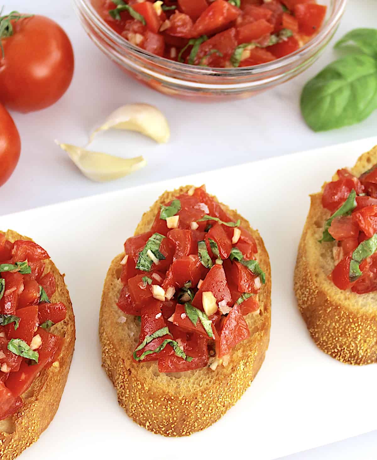 3 slices of Easy Bruschetta on white plate with tomatoes in background