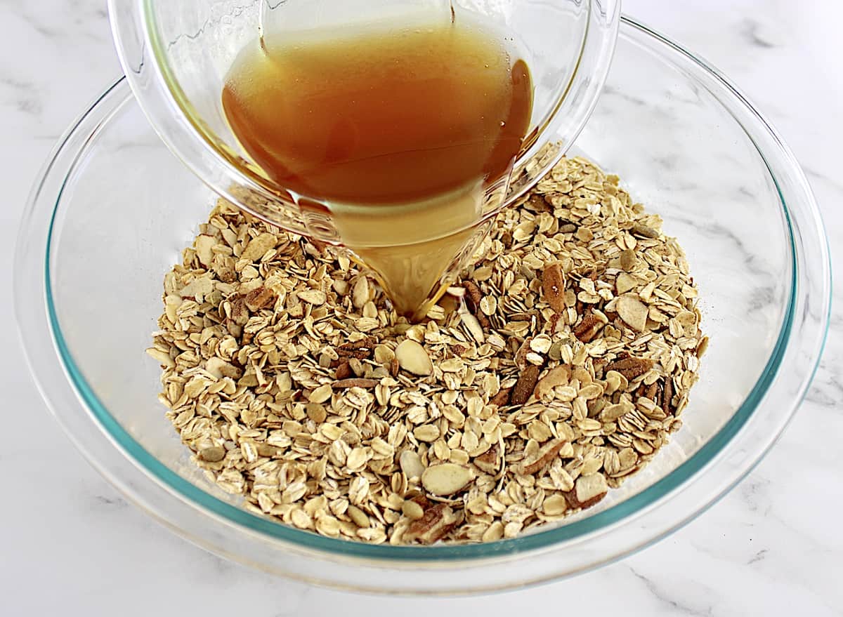honey and coconut oil being poured into granola mixture in glass bowl