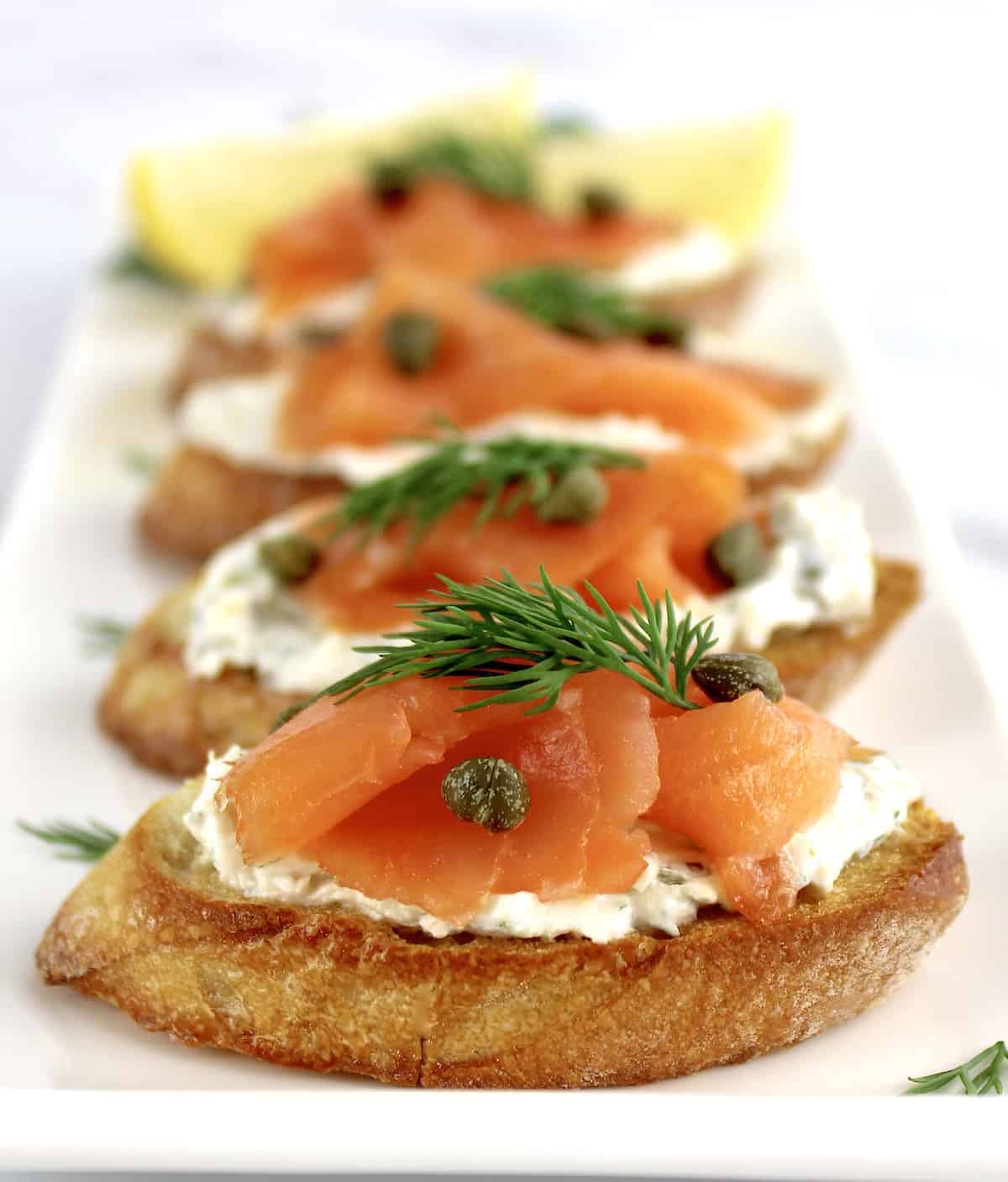 4 pieces of Smoked Salmon and Goat Cheese Crostini on white plate in a row