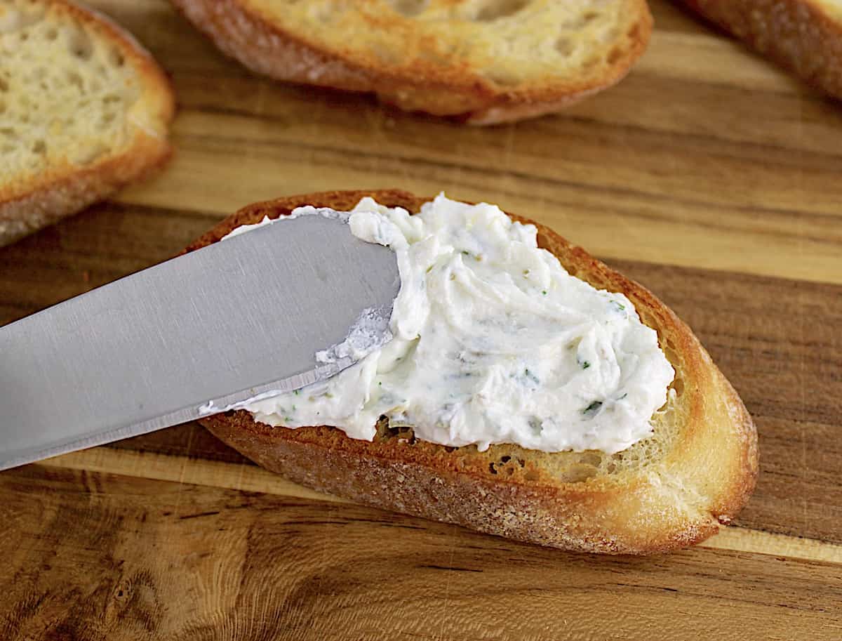goat cheese being spread on to toasted baguette slice on cutting board