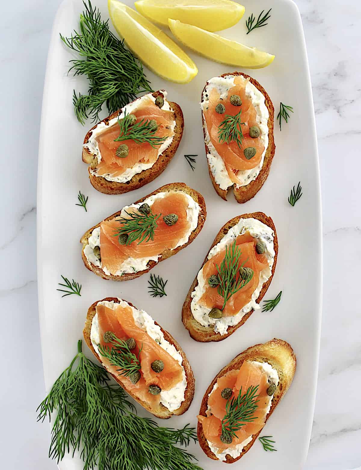 overhead view of Smoked Salmon and Goat Cheese Crostini on white plate with fresh dill and lemon wedges on side