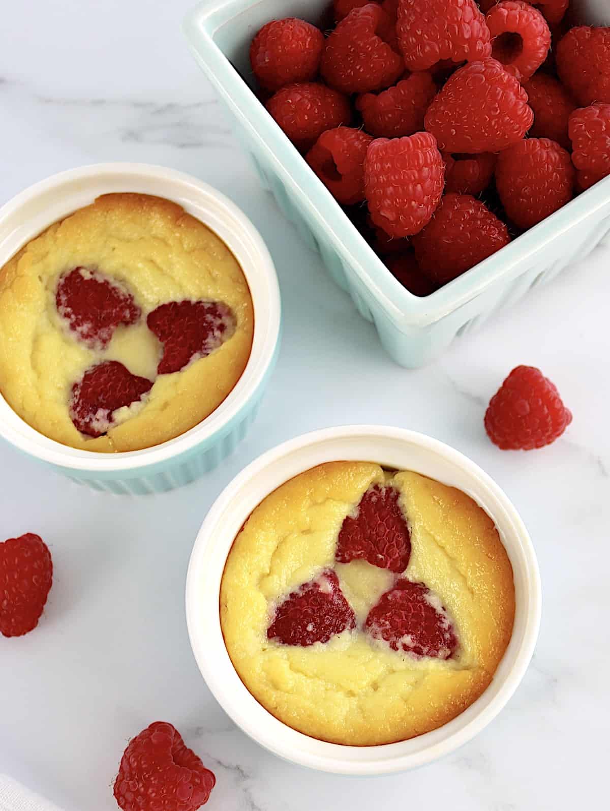 overhead view of 2 Baked Ricotta Raspberry Puddings with pint of raspberries in back
