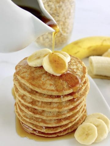 Banana Oat Flour Pancakes stack with sliced bananas on top and syrup being poured over top