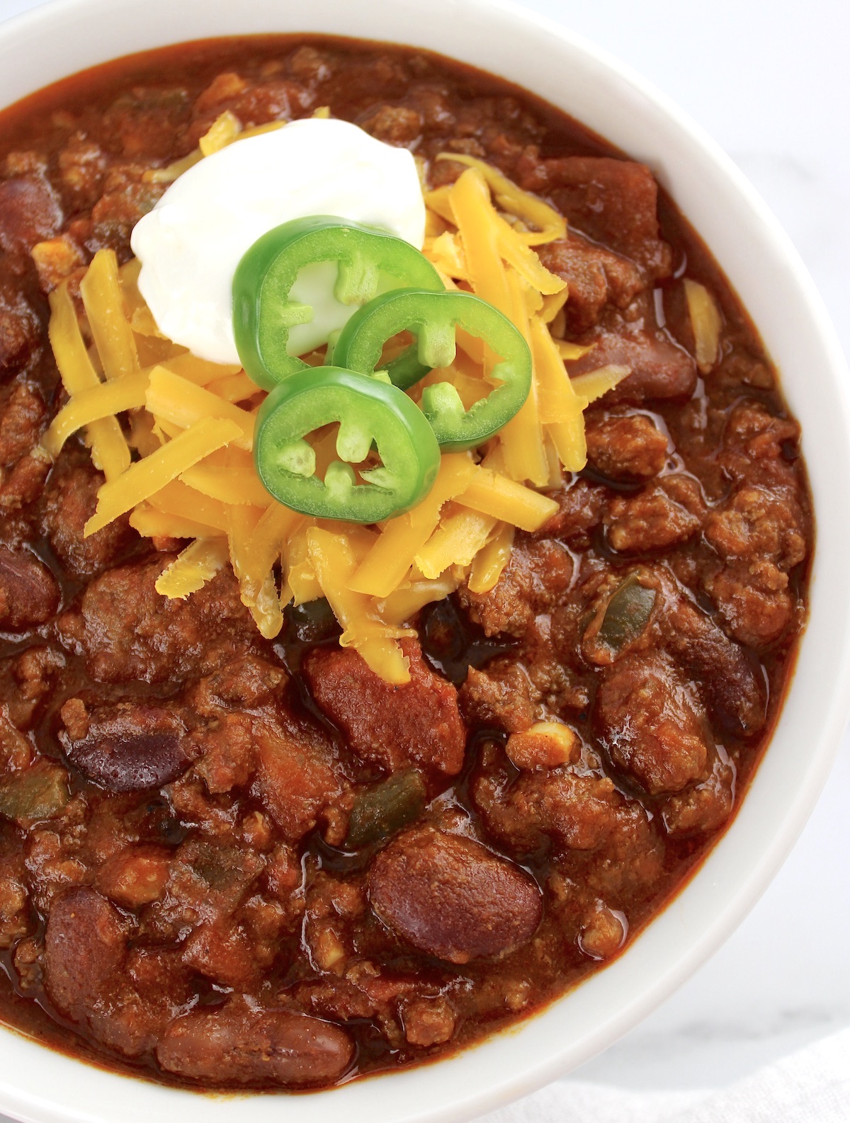 Best Homemade Chili in white bowl with shredded cheese, jalapeno slices and sour cream on top