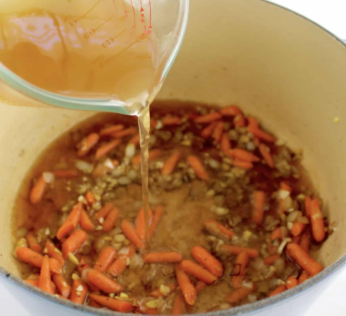 chicken broth being poured into pot with carrots and onions