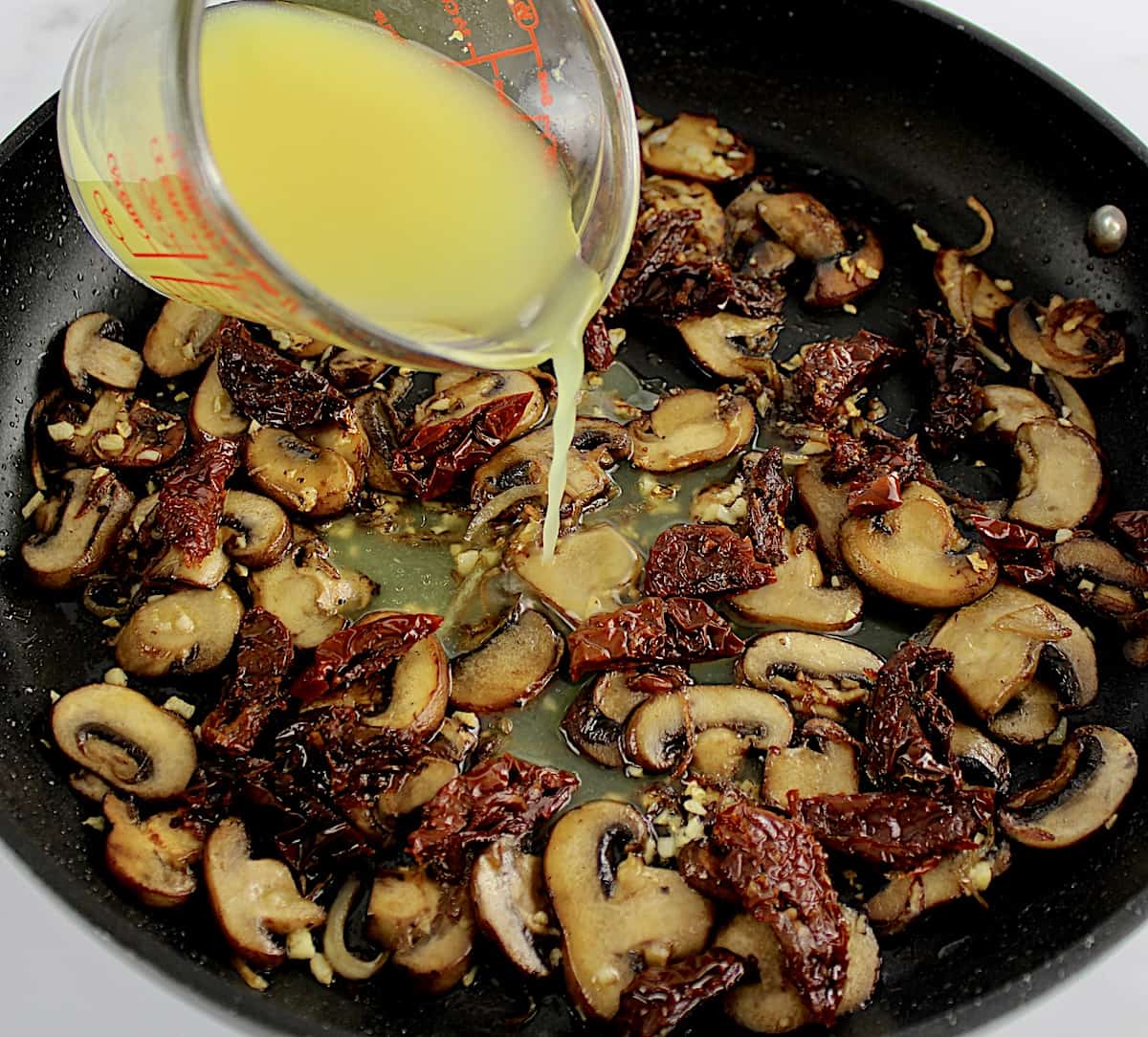 sauteed mushrooms, onions and sun-dried tomatoes in skillet with chicken broth being poured in