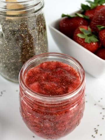Strawberry Chia Seed Jam in open glass jar with strawberries and chia seeds in background