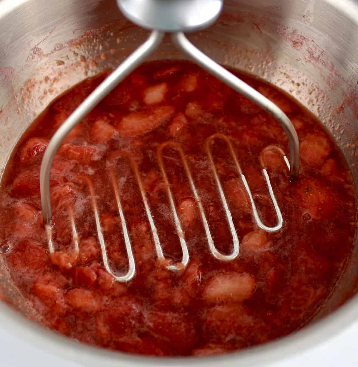 strawberries cooking in saucepan being mashed with potato masher