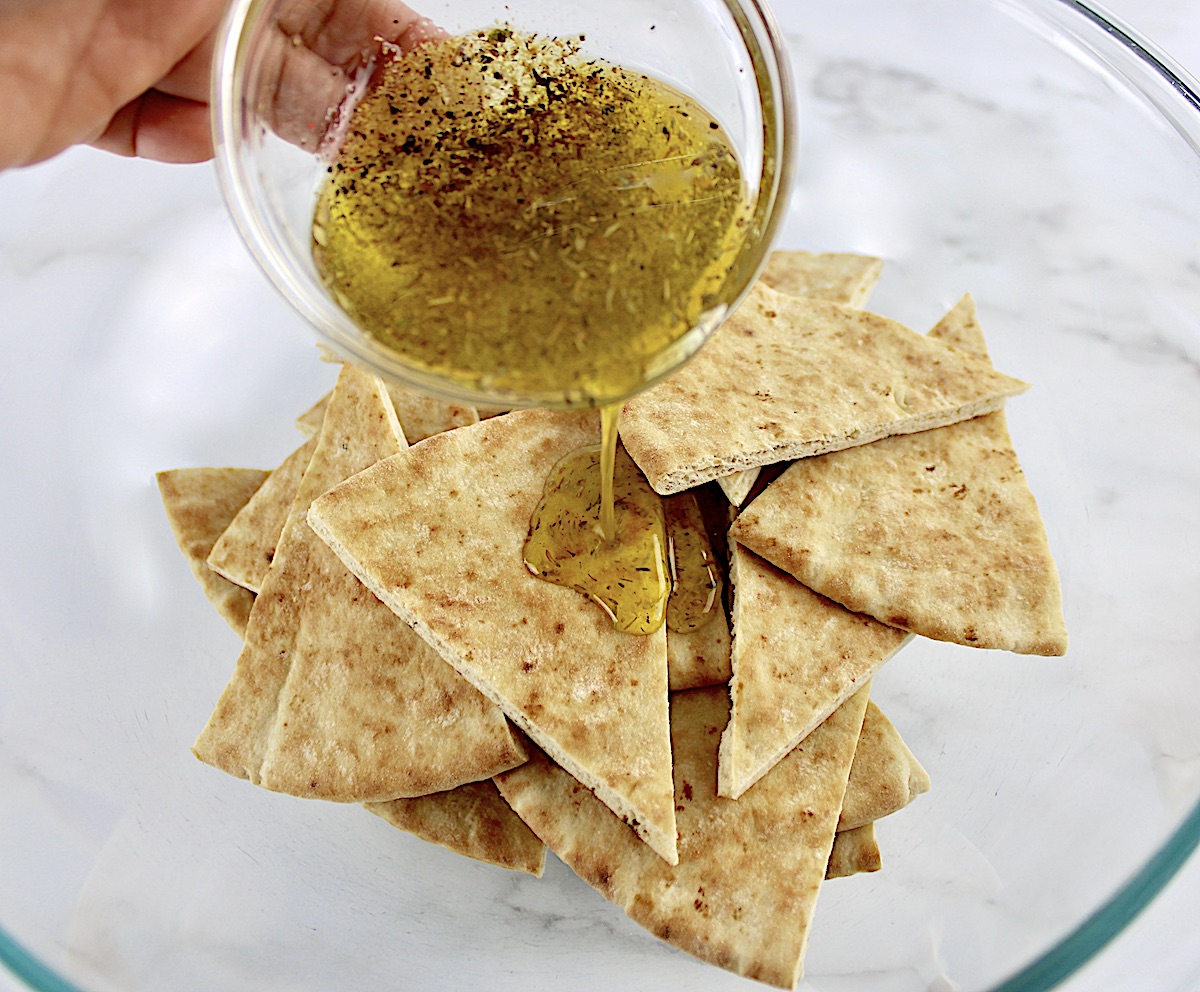 herbed olive oil being poured over pita triangles in glass bowl