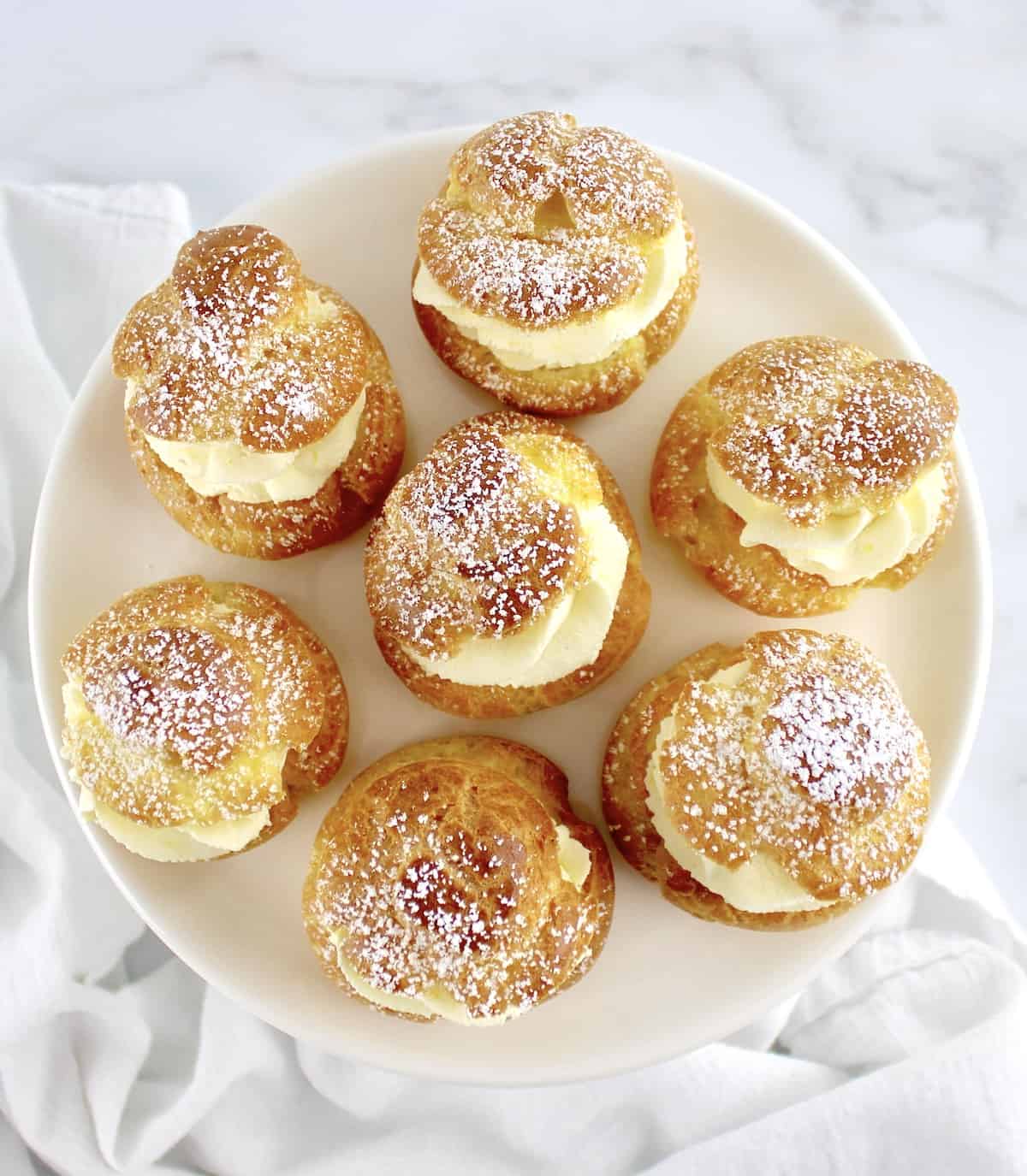 overhead view of 7 cream puffs on white round plate with powdered sugar on top